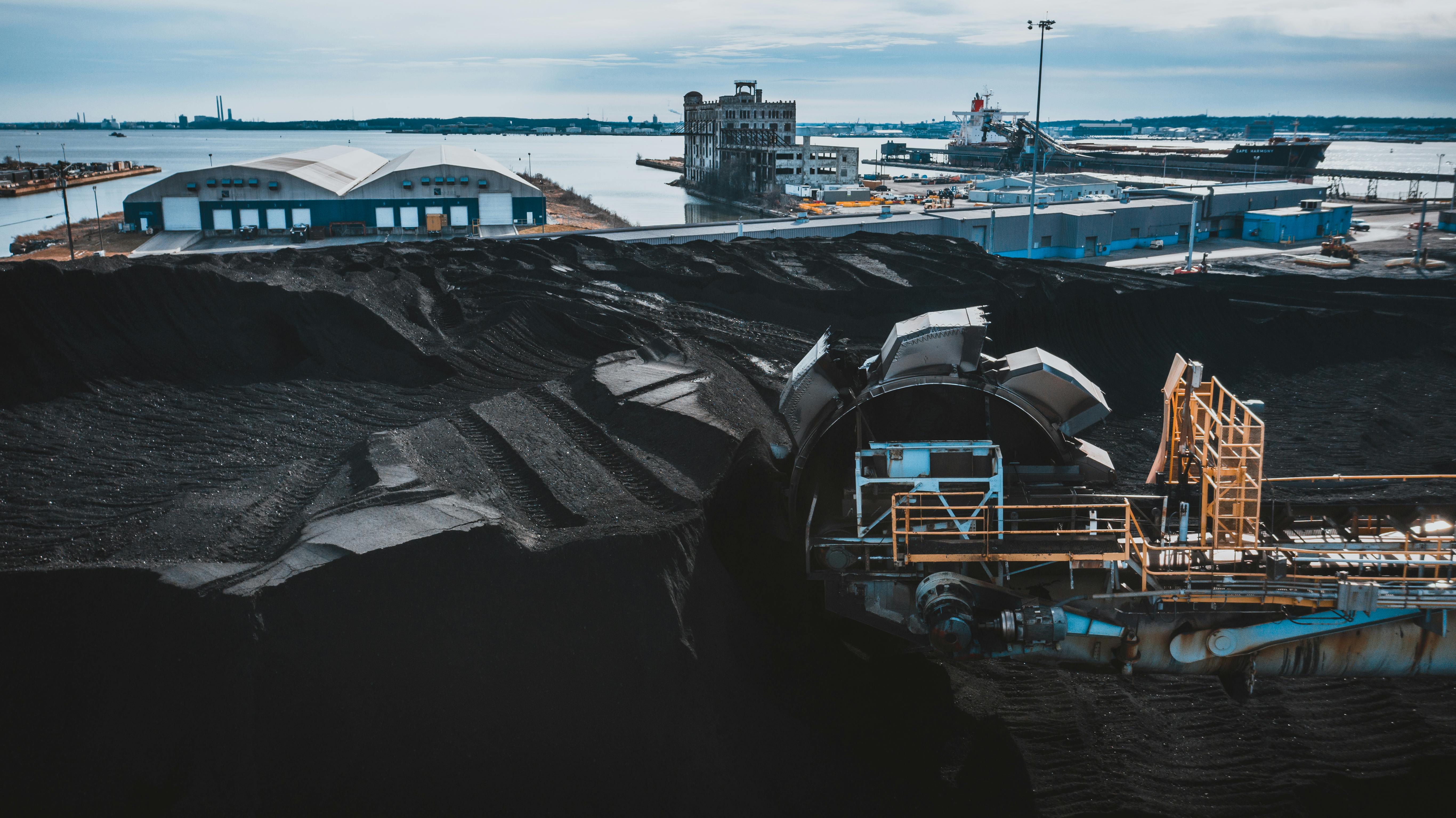 Large commodity stockyard beside port infrastructure with ship, storage sheds, and reclaiming equipment